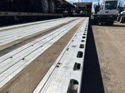 Flatbed trailer deck with metal and wood planks showing hole cutouts, side edge, and wheel in the background.