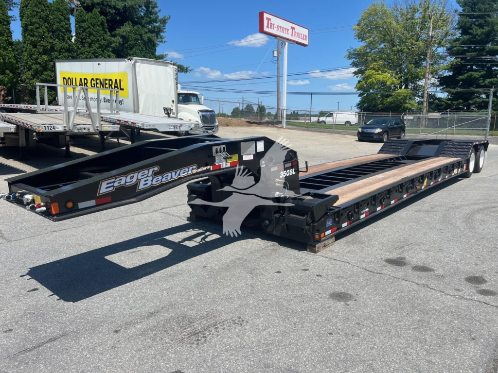 Lowboy trailer in a dealership lot showing Eager Beaver logo on the frame and long flatbed with multiple axles, and a white cab-over trucks in the background.