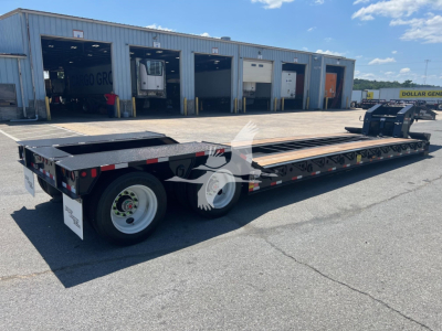 Lowboy trailer with long flatbed on site showing dual rear axles and extendable ramp area, VL088762 stock tag visible, warehouse bays in background.