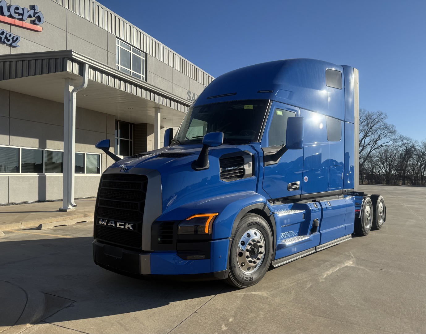 Blue Mack PR64T76H conventional sleeper truck in front of dealership building, showing cab with large windscreen, side mirrors, and dual rear axles.
