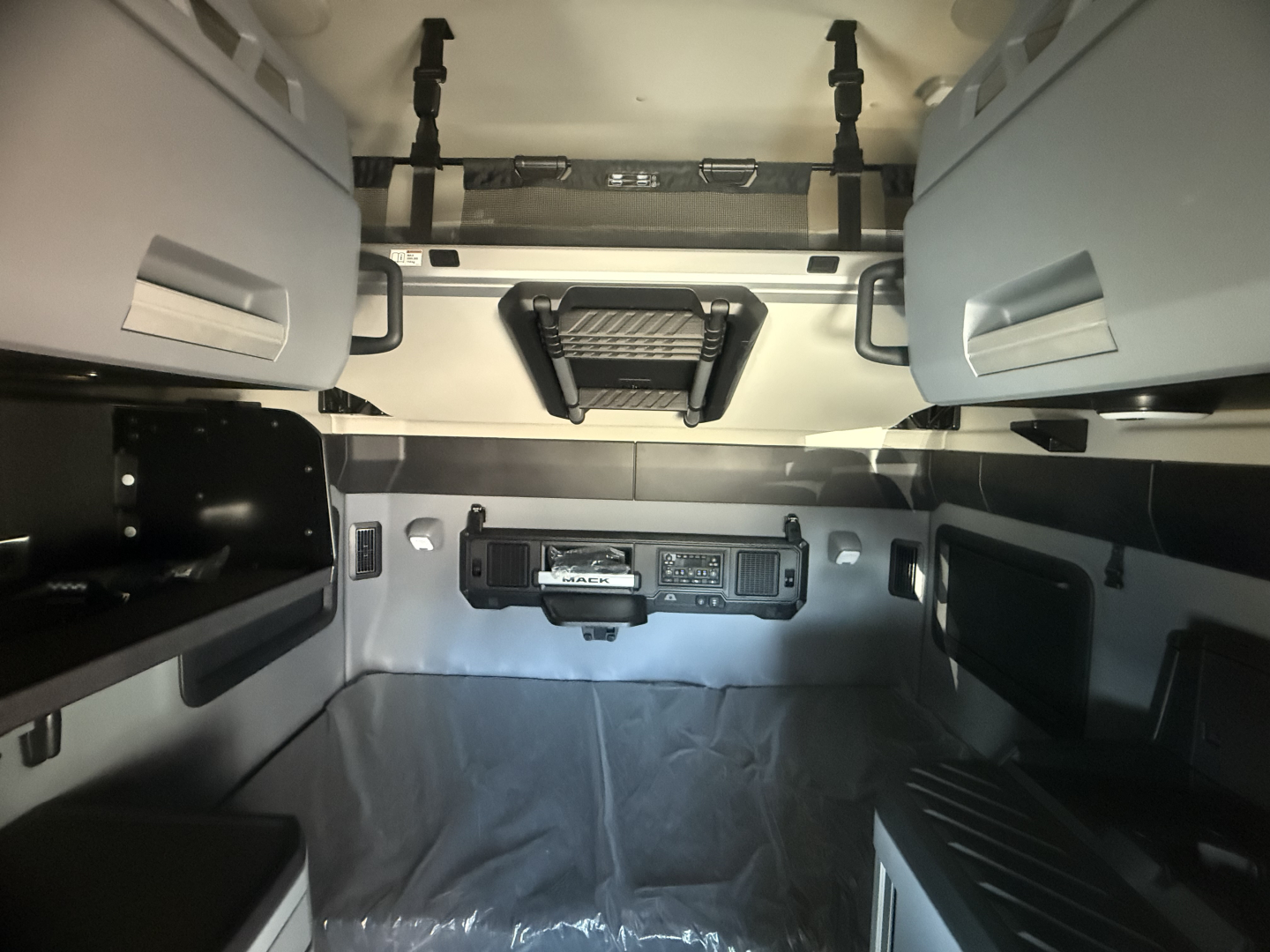 Cab interior of a new 2026 Mack PR64T76H sleeper showing overhead berths, storage cabinets, and control panel on wall.