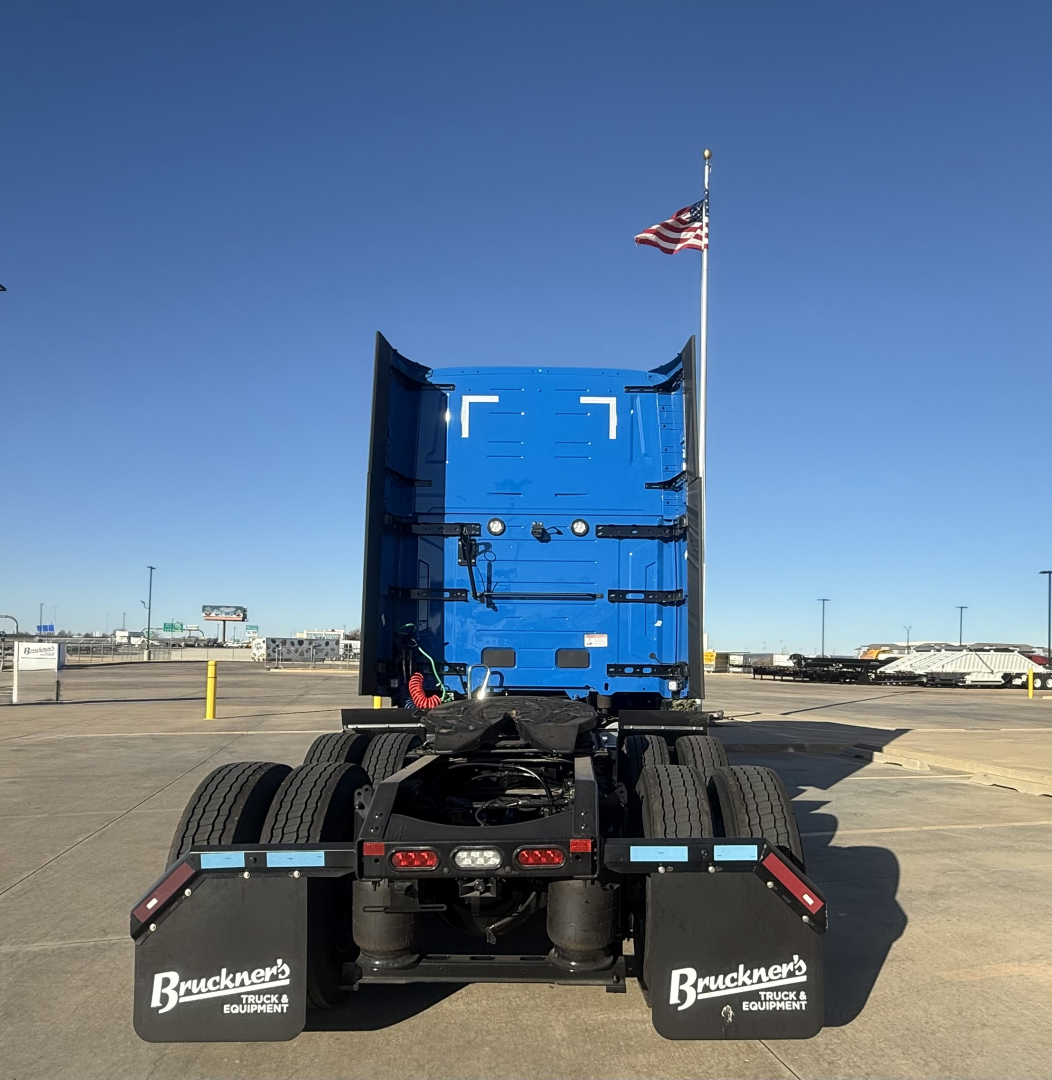 Truck rear view showing fifth wheel and dual rear axles on a blue Mack PR64T76H sleeper cab chassis. Visible tires, mud flaps with Bruckner’s Equipment logo, and US flag on antenna.