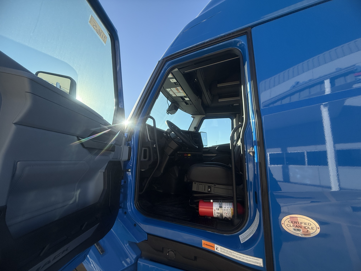 Truck cab interior of a blue Mack PR64T76H with open door showing steering wheel, dashboard, and a fire extinguisher mounted under the dashboard.