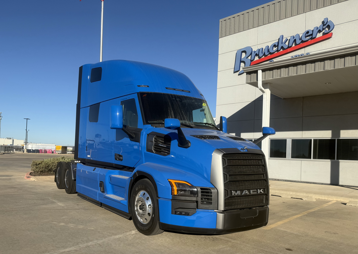 Blue Mack truck in front of dealership, PR64T76H model badge visible on grille area, large black front grille, side mirrors, and cab with sleeper height.