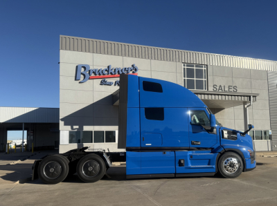 Truck cab in blue side view showing Mack PR64T76H wining details and dealership building in background.