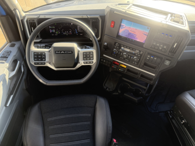 Truck cab interior showing steering wheel with Mack logo, dashboard with digital gauges and a center console with a screen. Visible labels include various control panels and interface elements.