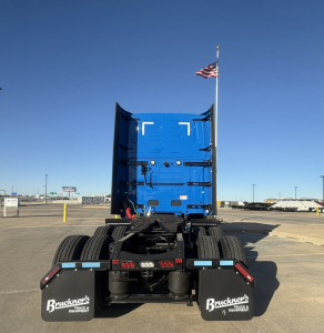 Truck rear view showing fifth wheel and dual rear axles on a blue Mack PR64T76H sleeper cab chassis. Visible tires, mud flaps with Bruckner’s Equipment logo, and US flag on antenna.