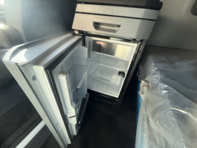 Mack truck cab interior with open under-seat refrigerator and small freezer compartment showing shelves and door bins.