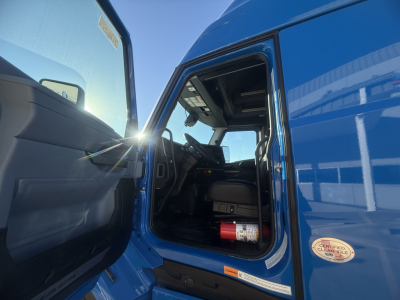 Truck cab interior of a blue Mack PR64T76H with open door showing steering wheel, dashboard, and a fire extinguisher mounted under the dashboard.
