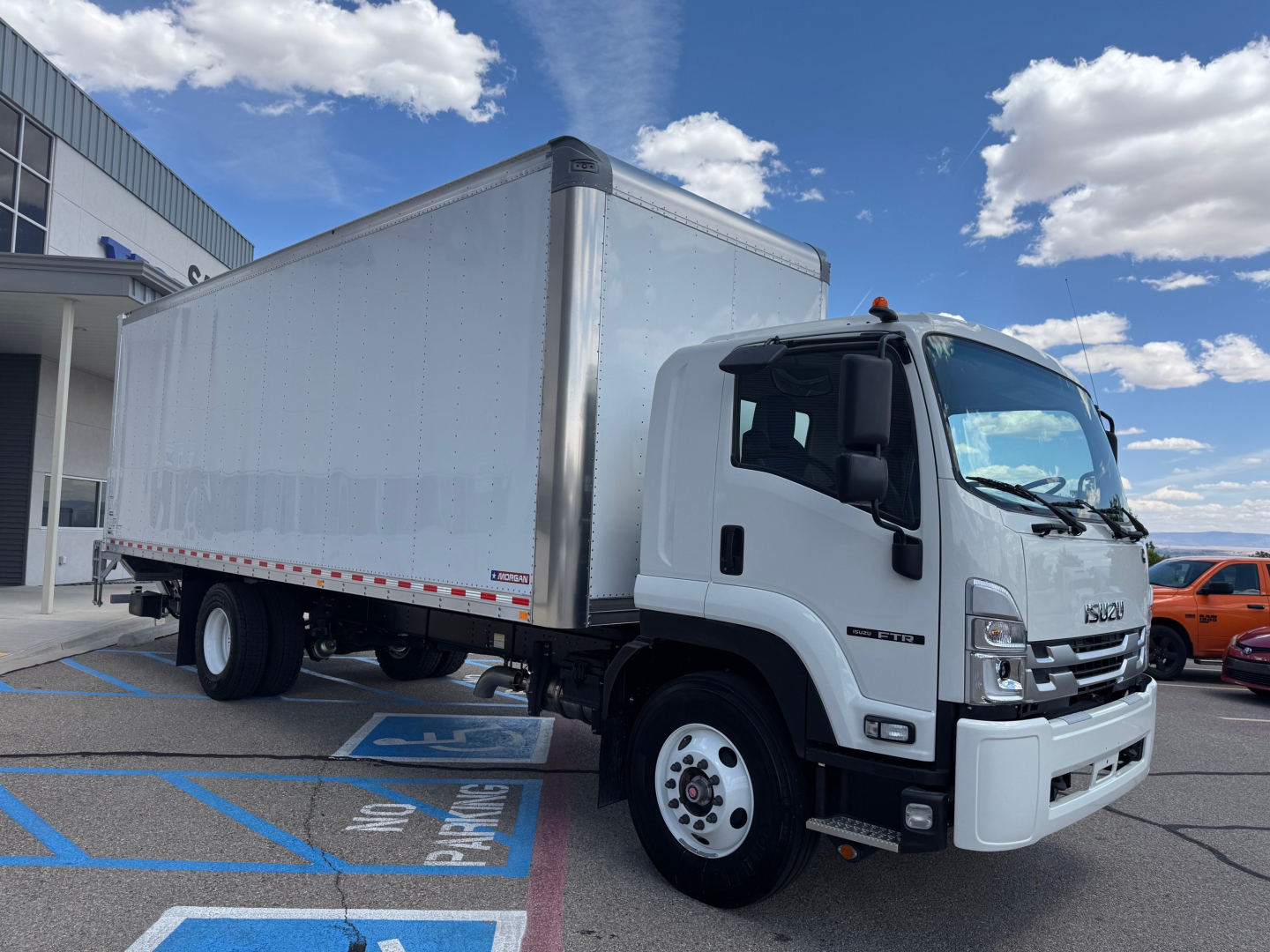 Isuzu FTR box truck in showroom lot, front-right view, white cab with large silver box body, single rear axle, clear blue sky.