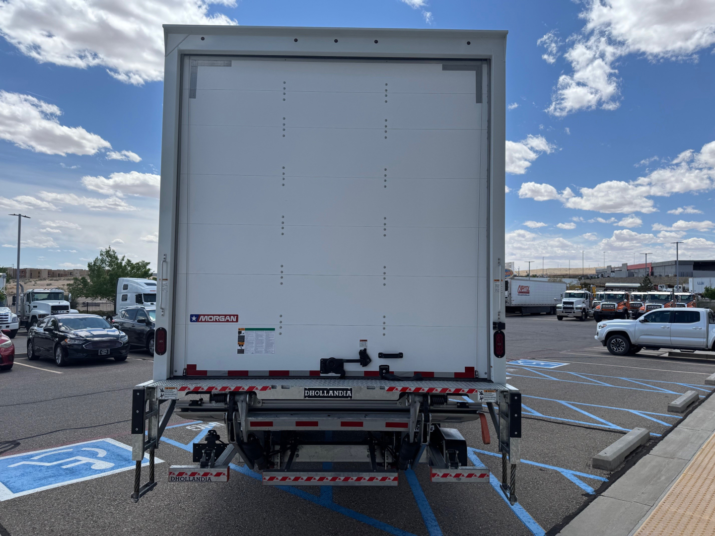Isuzu FTR truck rear view showing box cargo area and liftgate in up position, with bumper and taillights visible; blue sky with clouds in background.