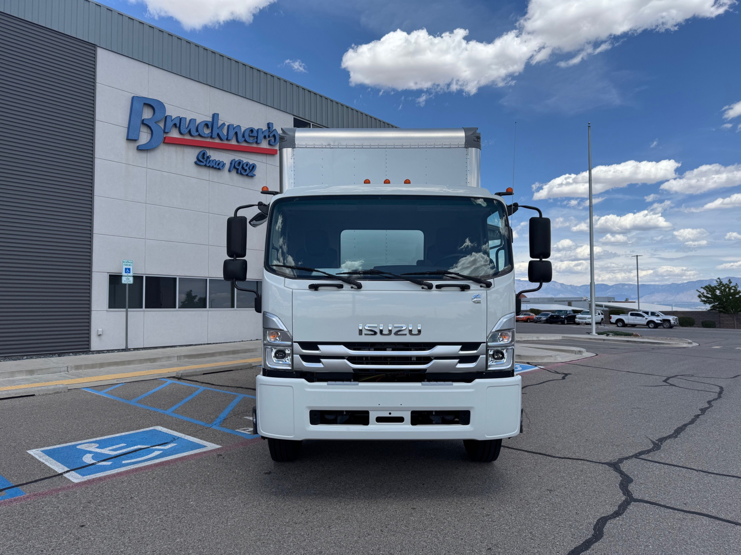 Isuzu FTR box truck front view in dealership lot with Bruckner's building in background and clear blue sky.