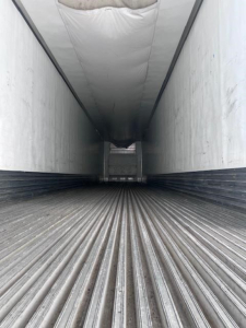 Interior of a trailer cargo space with corrugated floor and white sidewalls. Front landing gear area visible in distance.