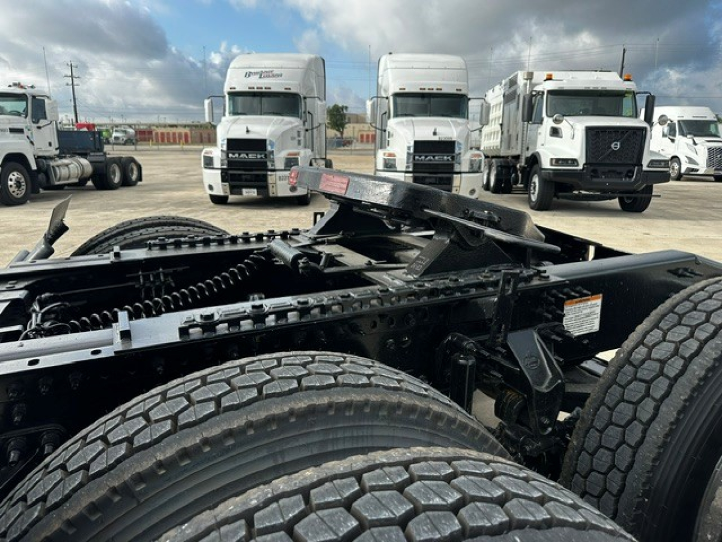 Truck chassis view showing dual rear axles and fuel/tank area under cab; '2019 Mack PI64T' listing context present in image seating area; front of several white trucks in background.