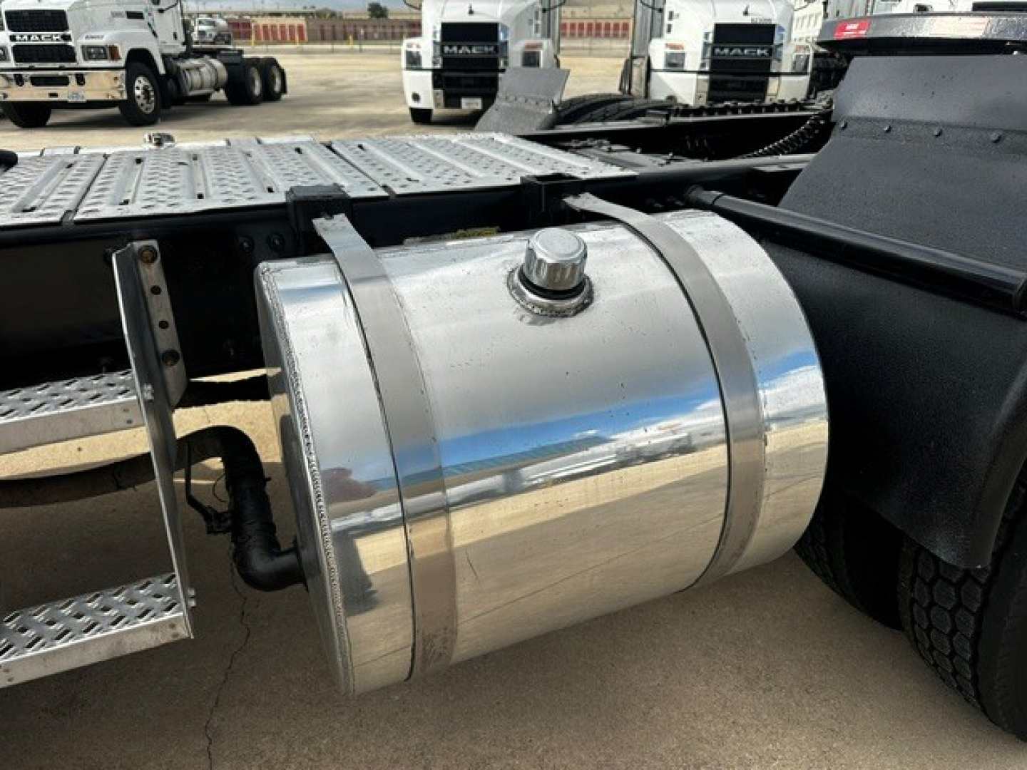 Fuel tank on a 2019 Mack PI64T tractor-trailer showing a polished cylindrical aluminum tank with cap and mounting strap, with other trucks in the background.