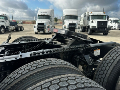 Truck chassis view showing dual rear axles and fuel/tank area under cab; '2019 Mack PI64T' listing context present in image seating area; front of several white trucks in background.