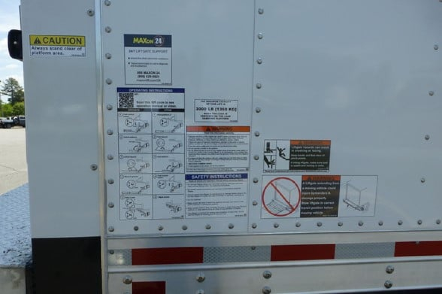 Truck trailer side with multiple safety and instruction stickers, including CAUTION label and MAXON 24 diagrams; small text panels and hazard icons.