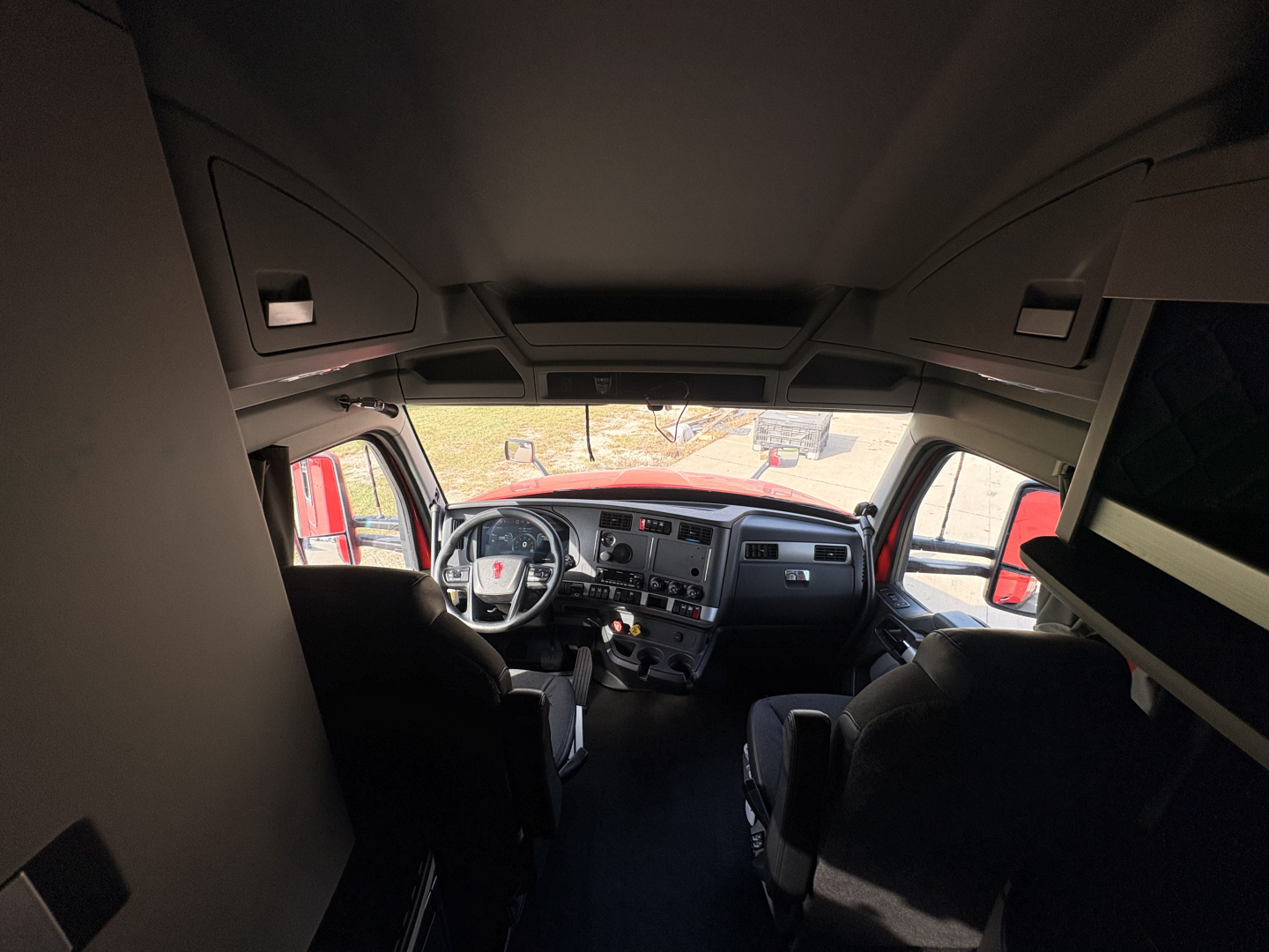 Kenworth T680 truck interior cab view from back of driver and passenger seats showing dashboard and steering wheel, front windshield, and overhead cabinets.
