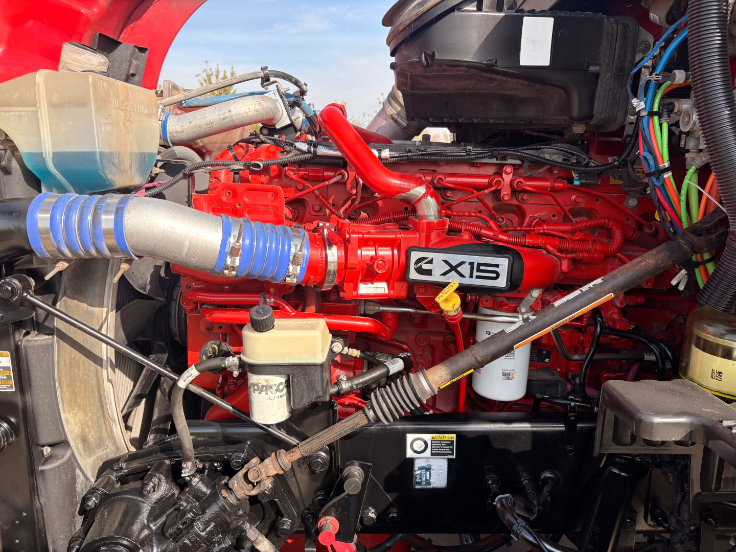 Kenworth T680 engine bay showing red Cummins X15 engine, blue turbo hose, coolant reservoir, and various hoses and wiring in engine compartment.