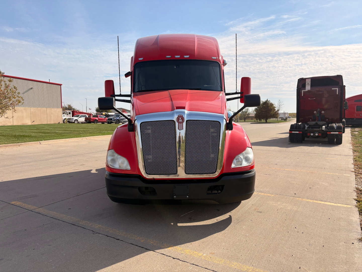 Kenworth T680 front view showing bold chrome grille and red cab with black bumper, large mirrors, and clear windshield; utility truck in background.