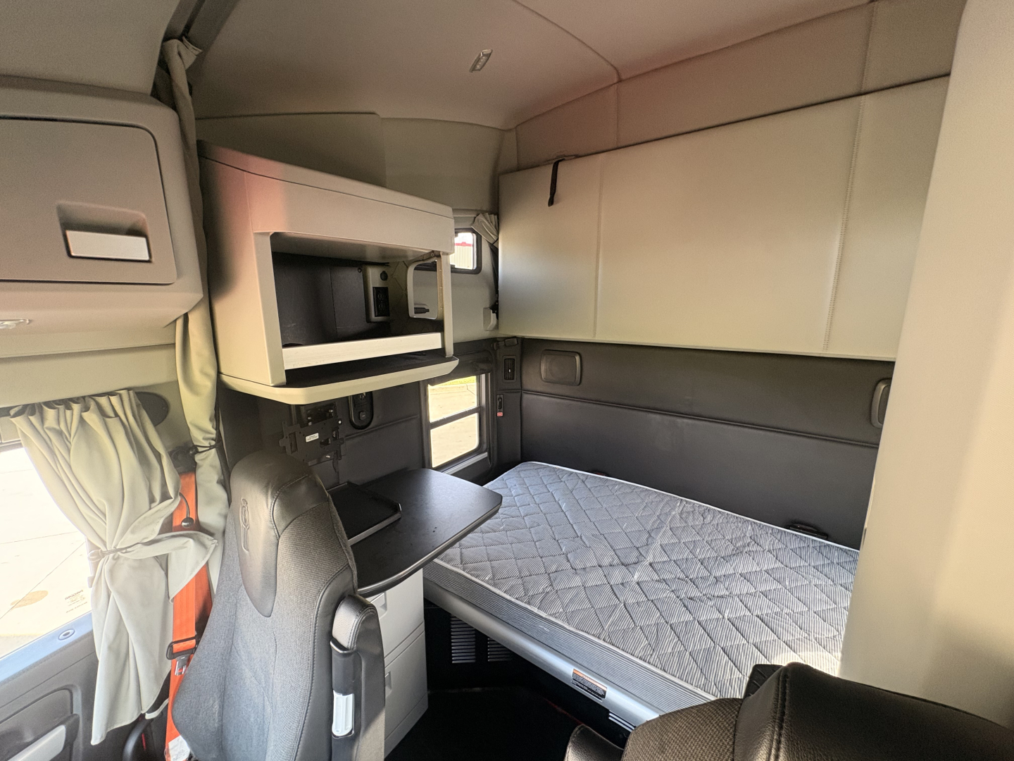 Interior of a Kenworth T680 sleeper cab showing a bed with quilted mattress, upper bunk shelf, microwave, cabinet, and desk area.