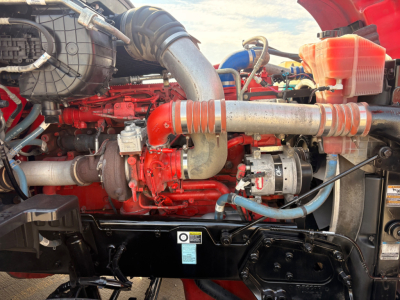 Kenworth T680 engine bay showing red engine block, intake piping, turbo, alternator, hoses, and coolant reservoir.