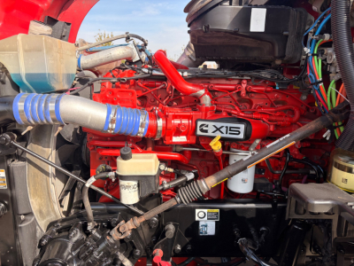 Kenworth T680 engine bay showing red Cummins X15 engine, blue turbo hose, coolant reservoir, and various hoses and wiring in engine compartment.