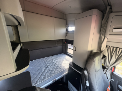 Cab interior of a 2022 Kenworth T680 sleeper truck showing a single bunk bed with quilted mattress, overhead cabinets, and a partition with a seat at the front.