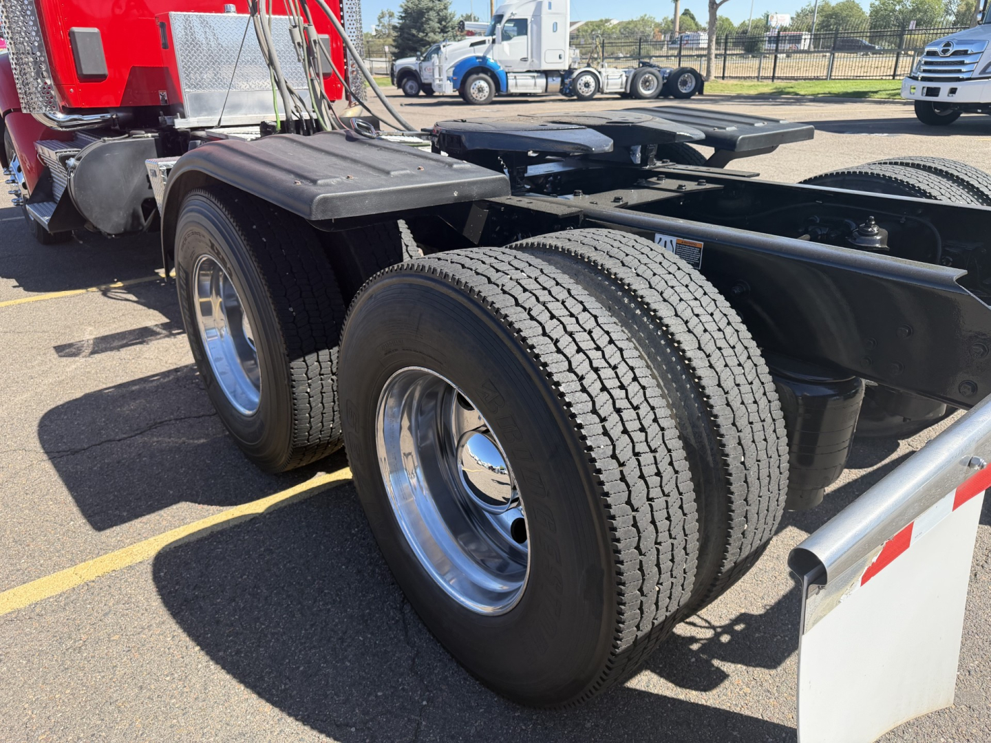 Kenworth T880 chassis with dual rear axles and tires in view; red cab in background; fuel tanks and frame visible; wheel rims chrome; no text readable on components.