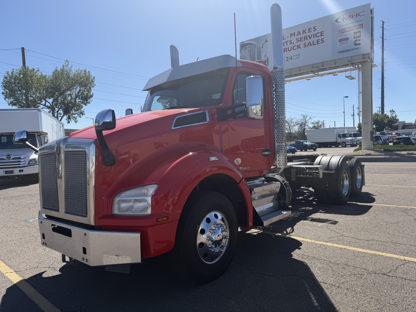 Kenworth T880 conventional daycab truck in red, front left view, with chrome bumper and dual rear wheels; visible side mirror; background signs and other trucks.