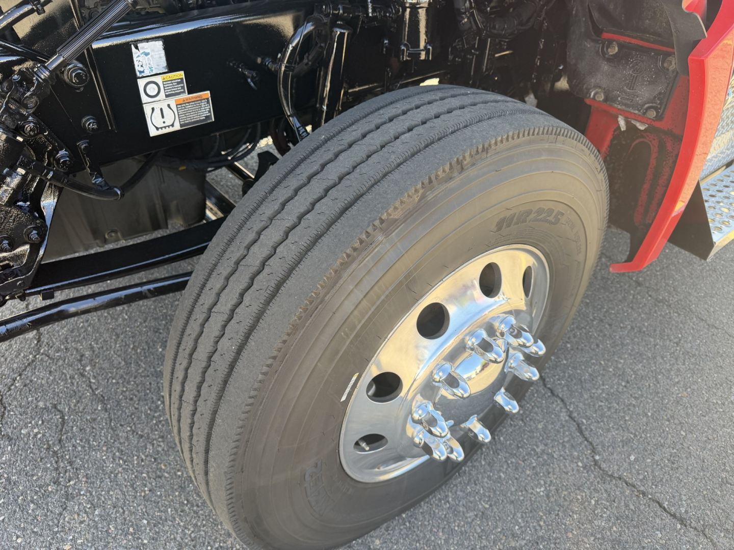 Kenworth T880 front wheel and suspension view showing dual rear steering? no, front axle with large chrome lug nuts and wheel cover; tire tread visible; part of truck frame and fender in view.
