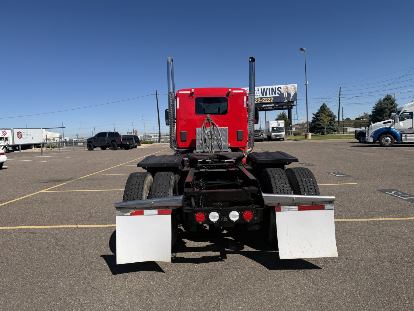 Kenworth truck rear view showing fifth wheel and dual rear axles in a truck lot; red sleeper cab with black frame and white mudflaps.
