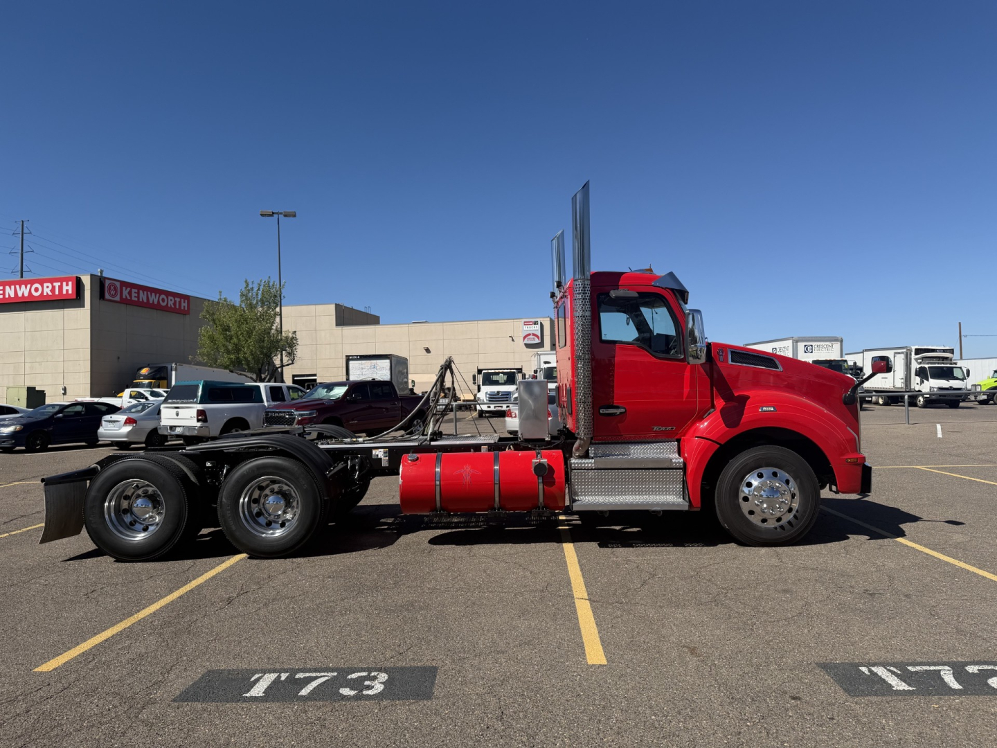 Kenworth T880 conventional daycab truck side view showing red cab, chassis with fuel tanks and dual rear axles, VIN plate not visible, parking lot lines.