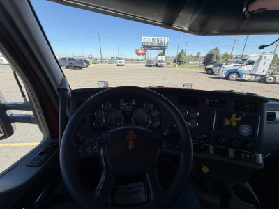 Kenworth T880 cockpit view showing steering wheel with Kenworth emblem, instrument cluster, dashboard with controls, and windshield view of truck lot.