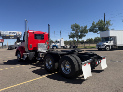 Kenworth T880 daycab tractor chassis in a parking lot, red cab, dual rear axles, with white mudflaps and chrome fuel tanks visible, no trailers attached.