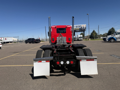 Kenworth truck rear view showing fifth wheel and dual rear axles in a truck lot; red sleeper cab with black frame and white mudflaps.