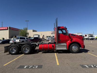 Kenworth T880 conventional daycab truck side view showing red cab, chassis with fuel tanks and dual rear axles, VIN plate not visible, parking lot lines.