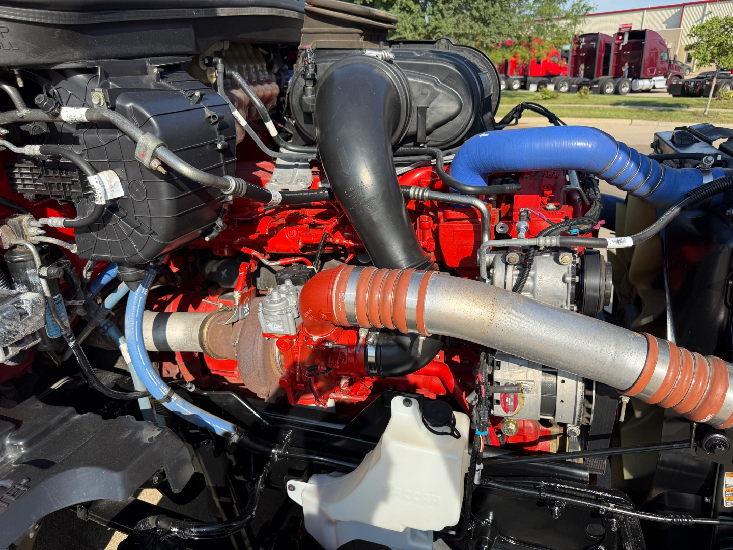 Kenworth T680 engine bay showing red engine block, blue silicone turbo hoses, and white reservoir; various hoses and metal components visible.