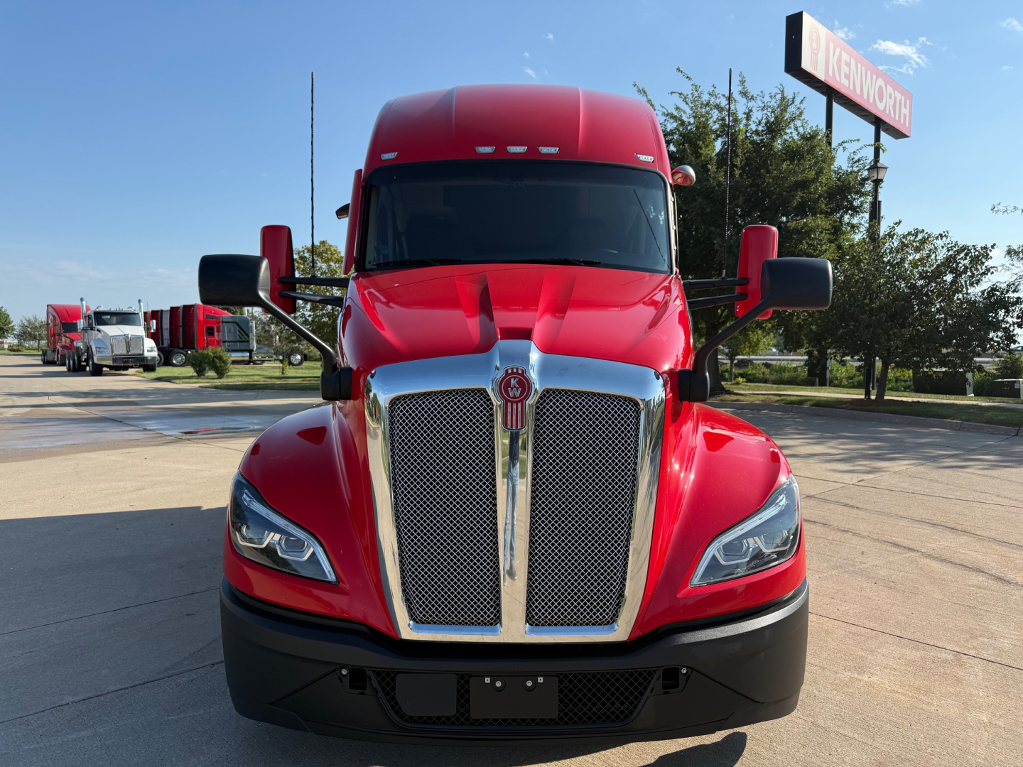 Kenworth T680 truck front view in red paint, chrome grille, black bumper, large side mirrors, daytime dealership lot in background.