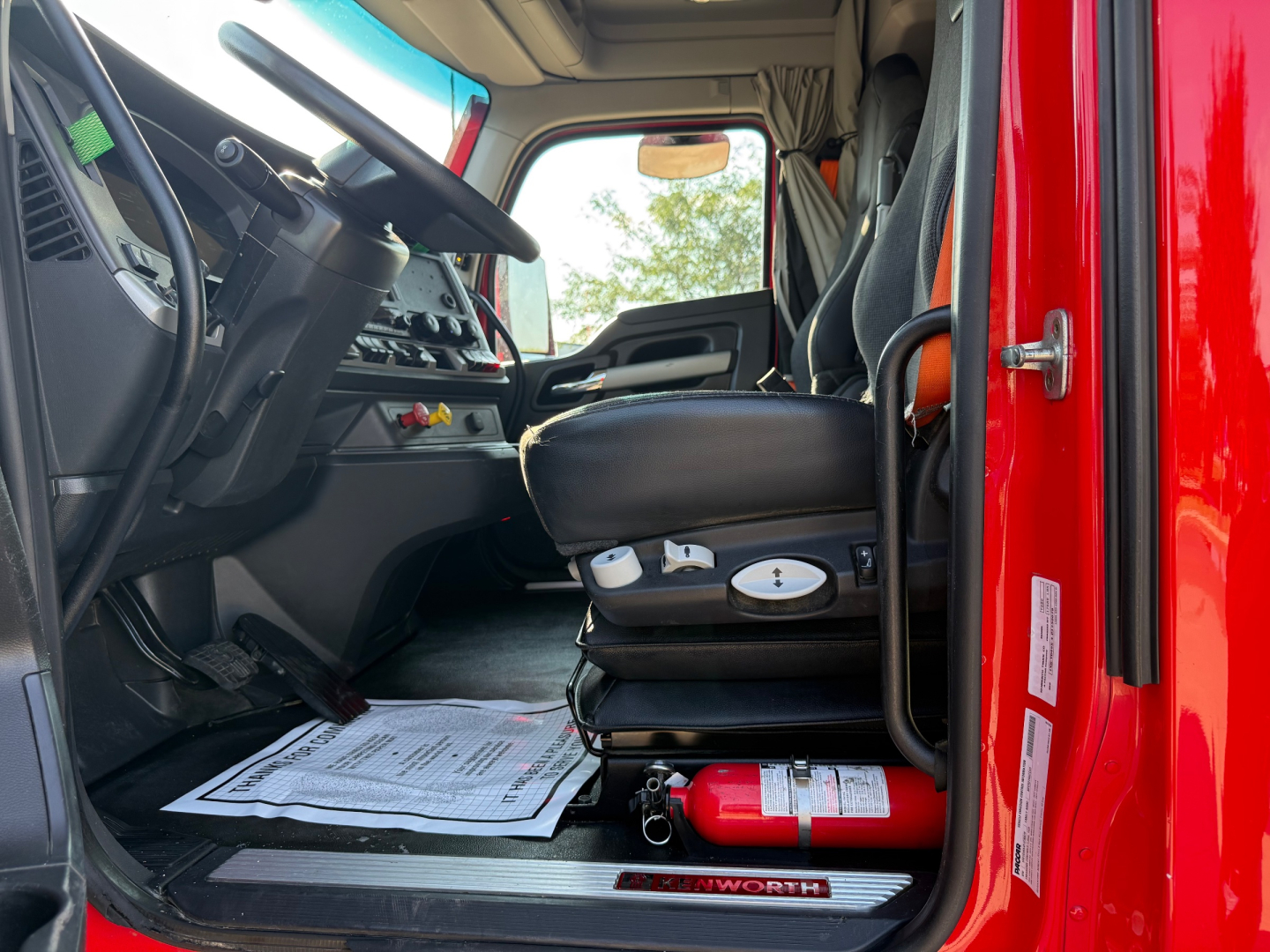 Kenworth T680 truck interior driver seat view with steering wheel, dashboard, and pedals; fire extinguisher visible under seat; red exterior visible at door edge.