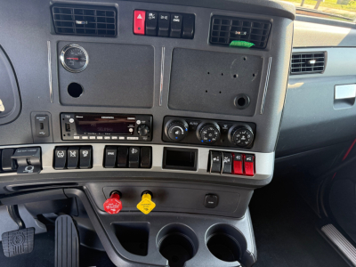 Cab dashboard of a Kenworth T680 with controls, gauge, and HVAC knobs visible; blank radio slot, two drink holders, and two colored knobs (red and yellow) near lower panel.