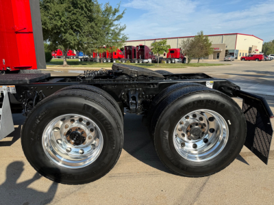 Kenworth T680 wheel area showing dual rear axles, chrome wheels, and part of chassis; outdoor dealership lot in background.