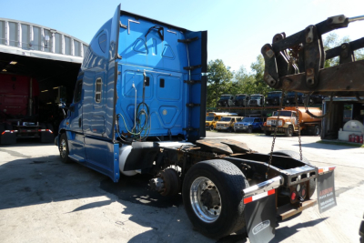 Truck cab and chassis of a blue 2014 Freightliner Cascadia 125 sleeper tractor viewed from the rear-left, with fifth wheel and hooks visible.
