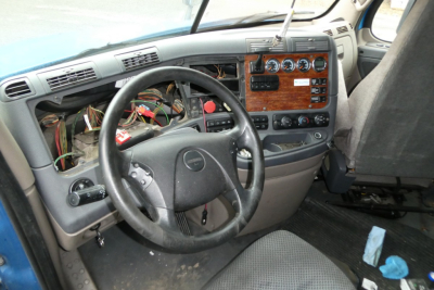 Cab interior of a Freightliner Cascadia truck showing steering wheel, exposed wiring, dashboard with woodgrain panels, and control dials.