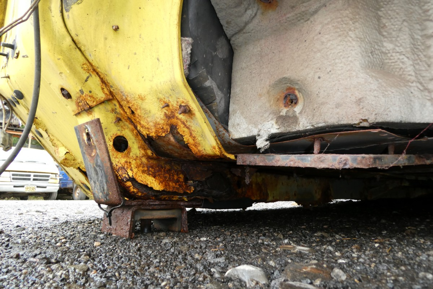Rear underside of yellow truck with rusted frame and support bracket visible near fuel tank; concrete ground.