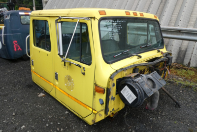 Front passenger side of a yellow International 4700 quad cab truck cab with exposed engine components, missing bumper, and visible day-coat labels on the windshield.