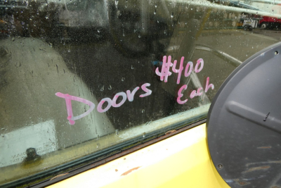 Truck window with pink handwritten text '#400 Each' and 'Doors' on glass; yellow vehicle body visible; circular door vent/hatch on right; dirt water streaks on window.