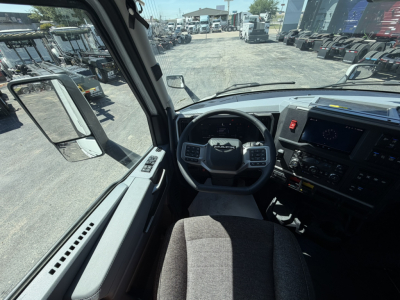 Cockpit view inside 2027 Mack PR64T76H showing steering wheel, dashboard, large touchscreen, and driver seat with side mirror; yard of trucks visible through windshield.