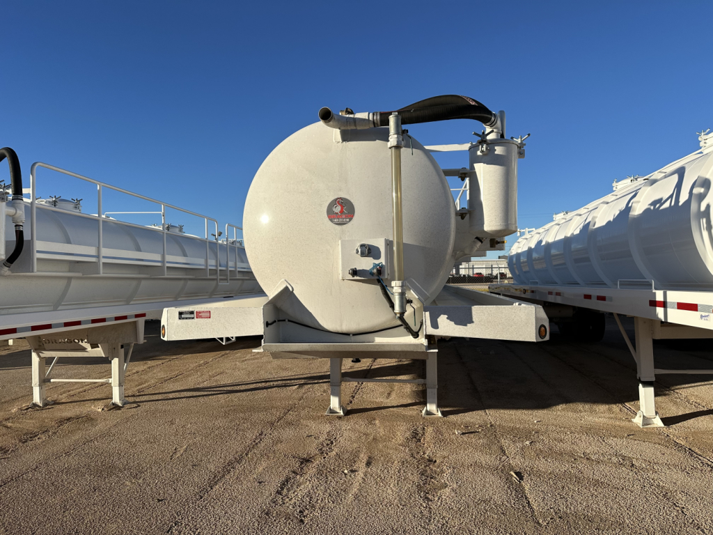  tank trailer with circular rear end and black hose, on a tri-axle chassis, with adjacent trailers on both sides and clear blue sky