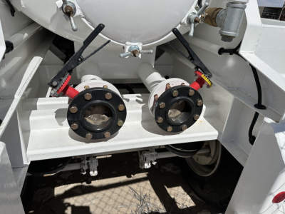 Two large flange fittings and valves on a white vacuum trailer chassis, dual circular flanges with bolts, hoses connected, visible undercarriage and frame.